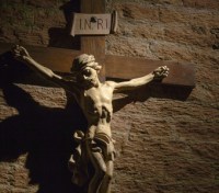 Dark artistic cross on a shaded rough textured brick wall, lit by a sun ray, with copy space for text